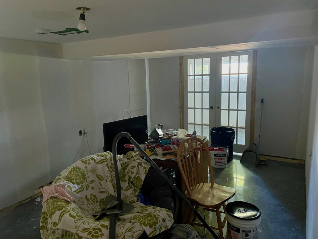 Sous-sol reconstruit après inondation avec sol foncé en époxy, murs blancs, mobilier temporaire et lumière limitée provenant de portes vitrées.